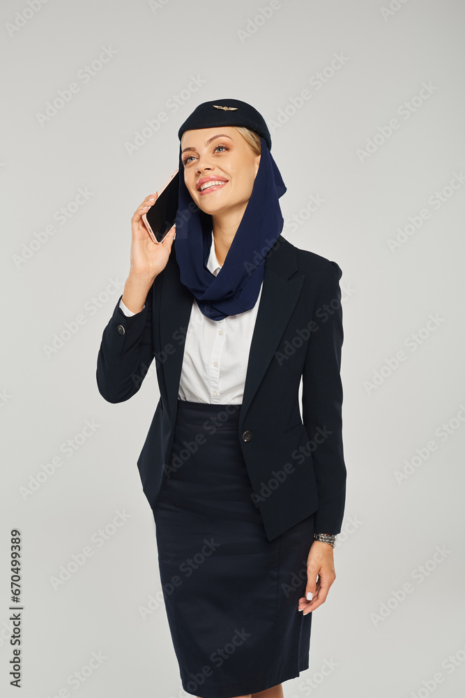 smiling stewardess in arabian airlines uniform talking on smartphone and looking away on grey smiling stewardess in arabian airlines uniform talking on smartphone and looking away on grey