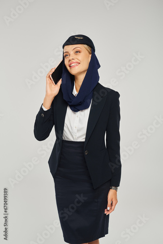 smiling stewardess in arabian airlines uniform talking on smartphone and looking away on grey