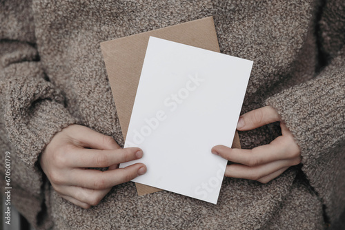 Winter, autumn cozy stationery still life. Child, female hands in beige pullower holding blank greeting card, craft envelope. Invitation mockup on knitted wool sweater. Blurred background. photo