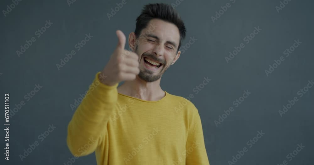 Slow motion portrait of happy young man showing thumbs-up hand gesture expressing admiration and approval on gray background. Emotion and people concept.