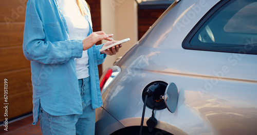 Close up of woman hands holding smartphone. Girl plugs power cable to charge electric car in parking near home. Eco friendly alternative energy green environment concept.