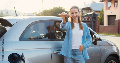 Happy adult woman showing keys standing on her new electric car outdoors on the street while charging. Girl boasts showing keys from recently purchased car.