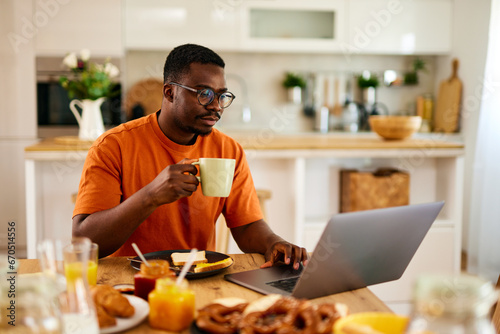 Pensive black man working on laptop during breakfast at home