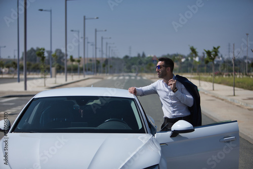 Successful, attractive, young Hispanic man, with white shirt and jacket over his shoulder and sunglasses, leaning on the roof of his white luxury car. Concept, cars, luxury, success, businessman. © Manuel