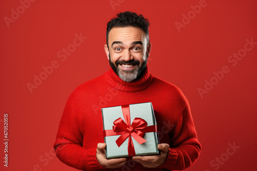 Happy fun man wearing with gift in ugly Christmas sweater on red background. Ugly sweater Day on December. Shopping and festive holiday sale. © svetlana_cherruty