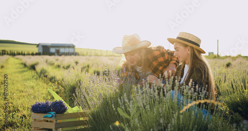 Cheerful and successful mother and daughter farmers wearing hats take care of lavender flowers on a green field at sunset. A woman and a girl are talking and working in the field. Small family