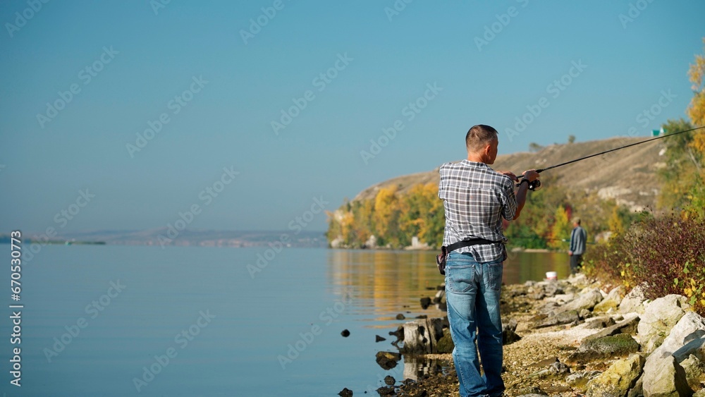A mature male fisherman in casual clothes casts a lure with a spinning rod while fishing on the river bank. Profile view of an adult man spinning fishing in the weekends. Copy space.