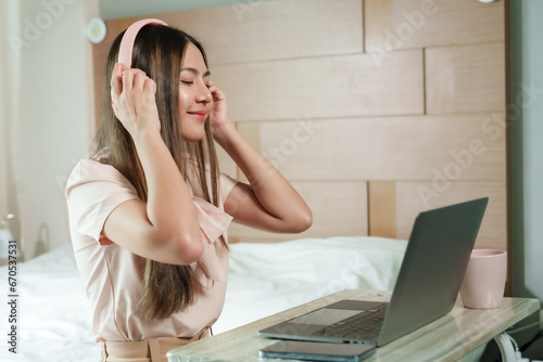 Beautiful Asian woman with long hair is wearing wireless headphones. Sitting happily listening to music and sing along, in front of a laptop computer on the bed in the bedroom Tired after working