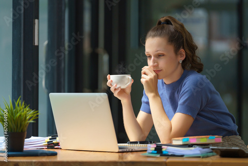 Beautiful businesswoman drinking coffee and using a laptop computer while working in an office.