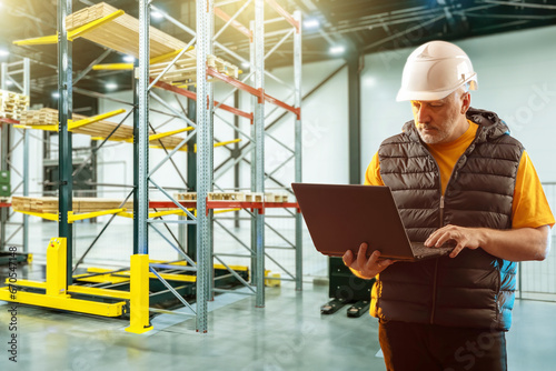 Man is storekeeper with laptop. Manager works in warehouse. Guy inside logistics center. Warehouse manager with computer. Man in hangar with warehouse racks. Contractor uses logistics software