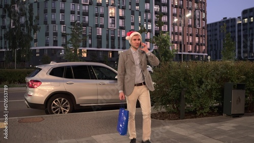 Happy young man in Santa Claus hat walking along the street, talking on mobile phone, car on the background. Business, celebration, New Year concept. Slow motion. © Nataliya