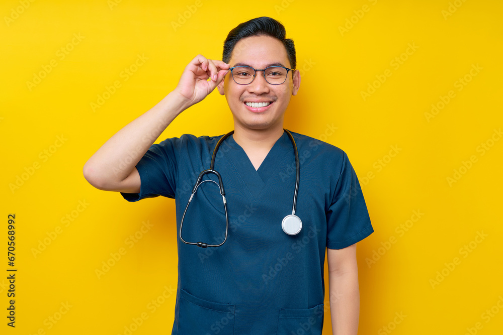Handsome young Asian man doctor or nurse wearing a blue uniform and glasses confident looking at the camera with smiling friendly isolated on yellow background. Healthcare medicine concept Handsome young Asian man doctor or nurse wearing a blue uniform and glasses confident looking at the camera with smiling friendly isolated on yellow background. Healthcare medicine concept