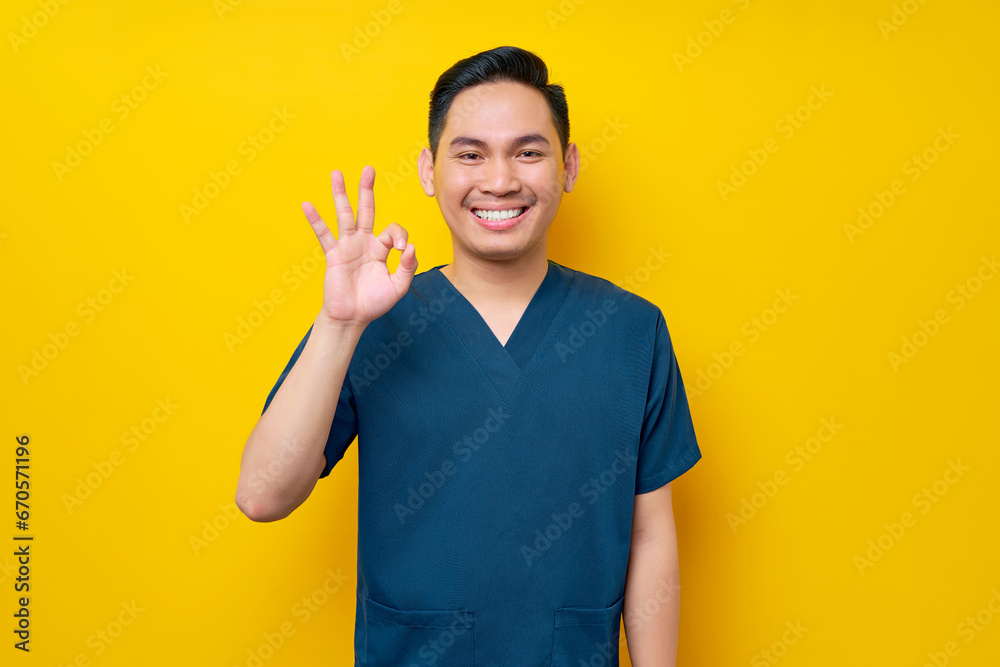 Professional young Asian male doctor or nurse wearing a blue uniform showing an okay sign, looking at camera with smile isolated on yellow background. Healthcare medicine concept Professional young Asian male doctor or nurse wearing a blue uniform showing an okay sign, looking at camera with smile isolated on yellow background. Healthcare medicine concept