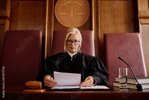 Canvas Print Confident mature female judge in black mantle looking through documents while si