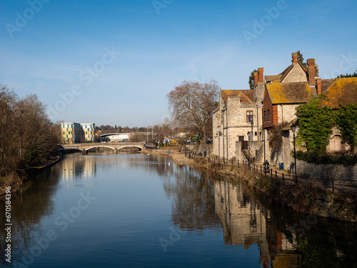 A north-west view, along the River Medway and paved riverside walkway, towards Maidstone Gyratory Bridge with period stone houses reflected in the water. Clear blue sky.