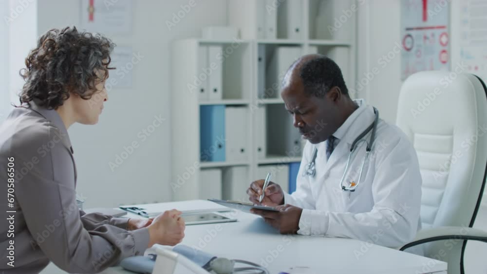 Medium side pan shot of black male practitioner talking to female Caucasian patient and writing down complaints in medical record in health clinic Medium side pan shot of black male practitioner talking to female Caucasian patient and writing down complaints in medical record in health clinic