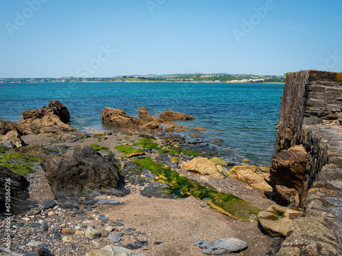 A shoreline of rocks, pebbles and gritty sand on the south Cornish coast at Polkerris. Bright blue sea. Distant hills. Clear blue sky. Space for text. photo