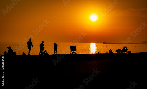A line of families and holidaymakers on the beach at Seaford  East Sussex  silhouetted in black against a red sky and setting sun.
