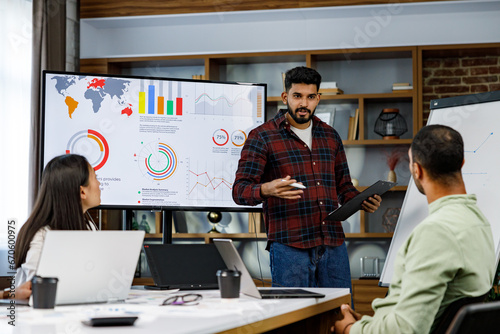 Motivated Indian guy team leader explains company development strategy to smiling young multinational colleagues. Happy multiracial business people enjoy working together in the office. Startup work.