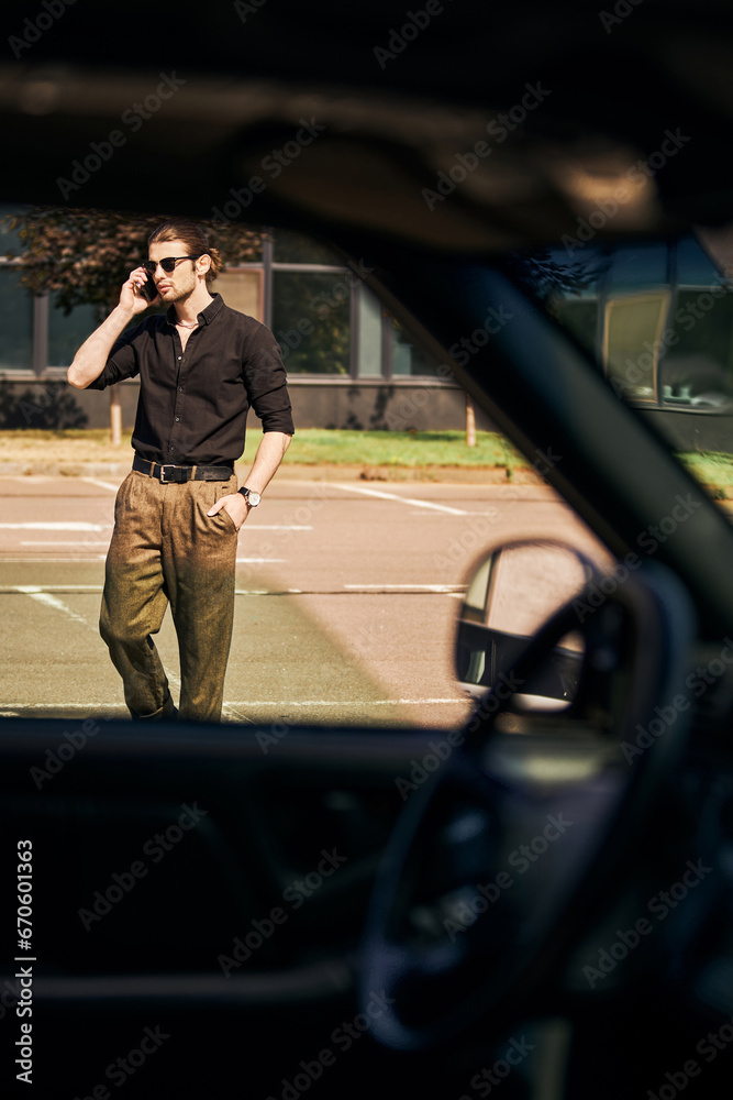 good looking man with dapper look with hand in pocket talking by phone with car insurer, sexy driver