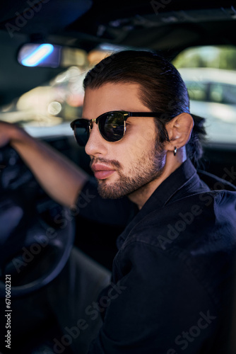 vertical shot of handsome sexy man in sunglasses in back shirt posing in his car, sexy driver