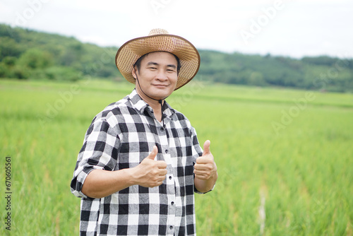 Handsome Asian man farmer is at paddy field, wears hat and plaid shirt, Two hands thumbs up. Happy and confident. Concept, agriculture occupation.  