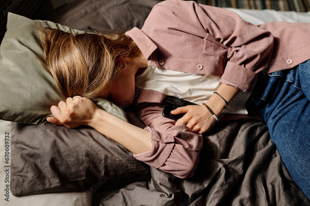 Frustrated teenager with smartphone in hand crying in pillow because of cyberbullying messages from anonymous people while lying in bed