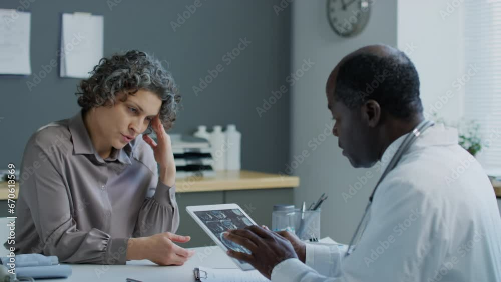 Medium pan shot of Caucasian woman with grey hair complaining about migraine to African American doctor who looking at MRI results on tablet in hospitalMedium pan shot of Caucasian woman with grey hai