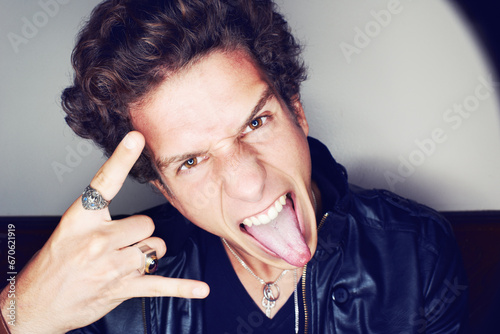 Rockstar man, horns and hand in studio for emoji in spotlight, fashion or leather jacket by background. Person, symbol and icon by halo for portrait, punk style and tongue with jewelry for aesthetic photo