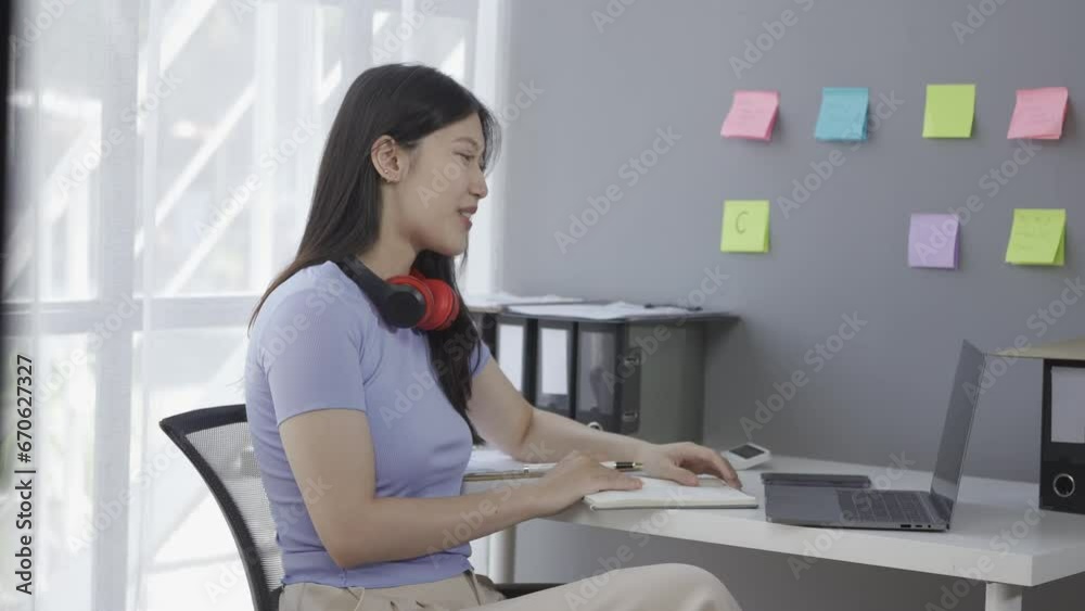 Young Asian female student wearing headphones surfs the internet on a laptop while taking an online class or lecture and doing a Google Zoom video call. Young Asian female student wearing headphones surfs the internet on a laptop while taking an online class or lecture and doing a Google Zoom video call.
