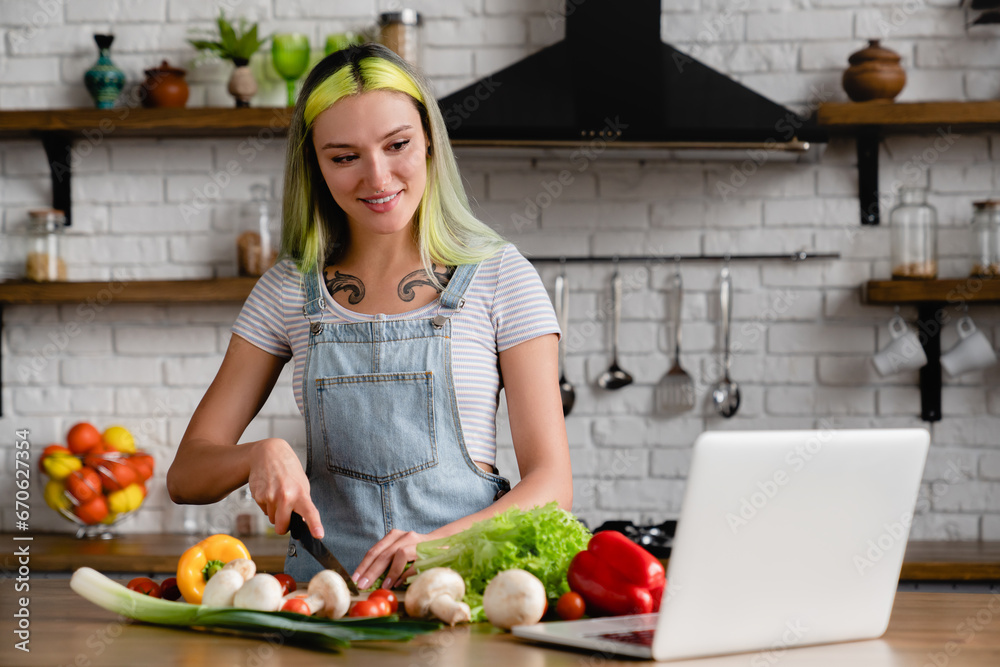 Happy diverse caucasian young teenage girl stylish woman with colorful hair tattoo cooking breakfast, preparing food, tasty dinner salad at home kitchen while using laptop with online tutorial recipe