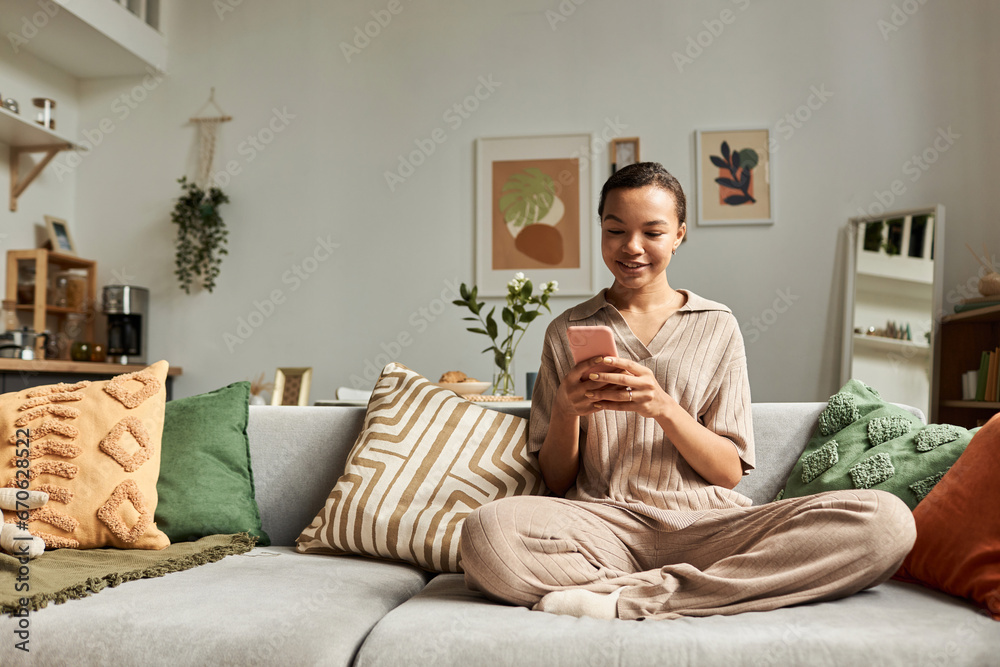 Front view portrait of smiling Black girl using smartphone while sitting cross legged on couch in cozy home, copy space Front view portrait of smiling Black girl using smartphone while sitting cross legged on couch in cozy home, copy space