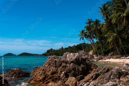 Blue sea and white sandy beach with tall coconut palm and rocks bright sky.