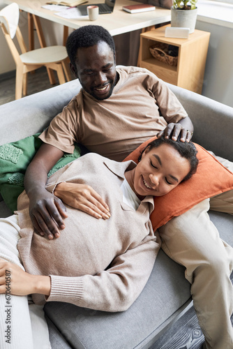 Vertical portrait of young Black couple expecting baby and cuddling on couch smiling happily