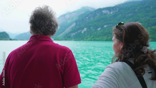 Back of Senior man and adult daughter traveling by boat observing lake and mountain view. Woman pointing at horizon exploring nature with father #670656566
