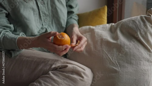 Close up medium shot of girl hands peeling mandarin orange fruit sitting on the floor near the window in cozy bedroom during rainy sad day