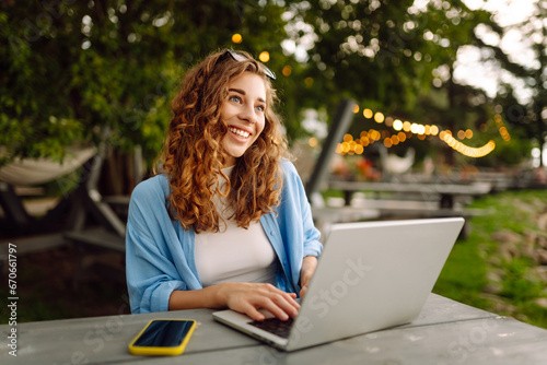 Happy woman in casual clothes working on a laptop in an outdoor cafe near a lake. Young woman freelancer enjoying nature. Tenology concept, working day.