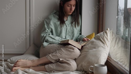 Attractive young female reading book turning pages sitting near the balcony window with pillows on the wooden floor at home in a sad day 