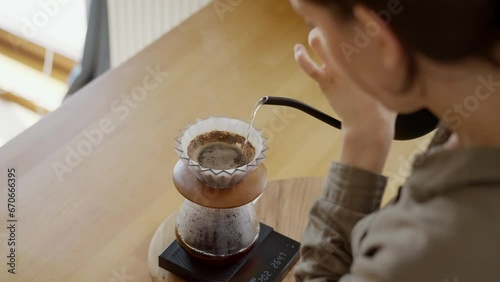 Young woman pouring hot water from kettle into v60 origami filter with fresh grinded coffee beans on wooden desk in the kitchen at home