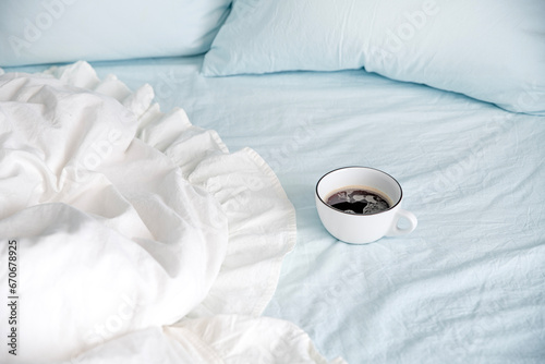 A white cup of coffee stands on a bed with pillows and a blanket with white linen and blue