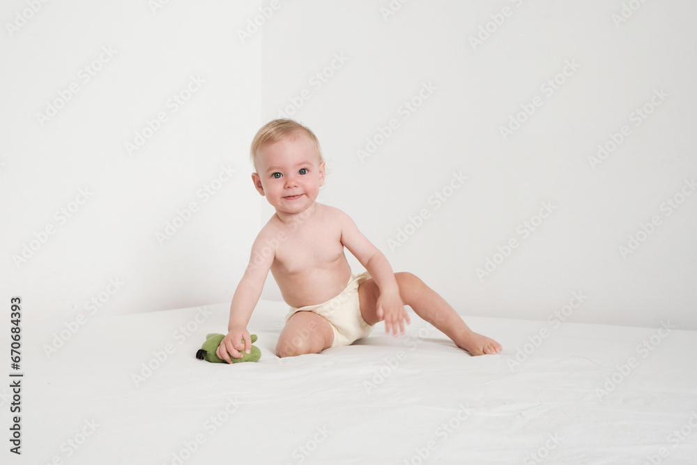 Happy baby in panties is playing with green plush toy on white bed in white room Happy baby in panties is playing with green plush toy on white bed in white room