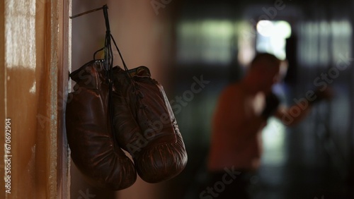 In the close up shot, brown leather boxing gloves hang on a cord from a rusty nail on a door frame. In the background, a man wearing boxing gloves stands in a stance. Training before the fight © kinomaster