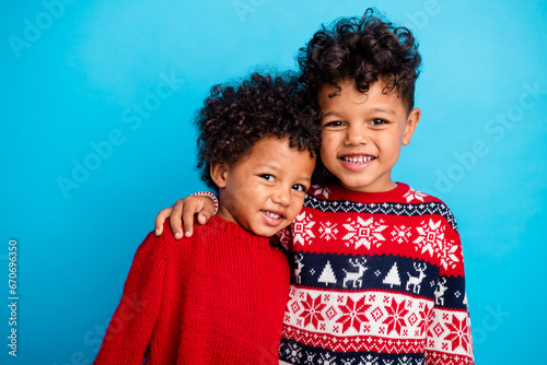 Portrait of two funny little friends with wavy hair dressed red ornament sweaters embrace on new year isolated on blue color background