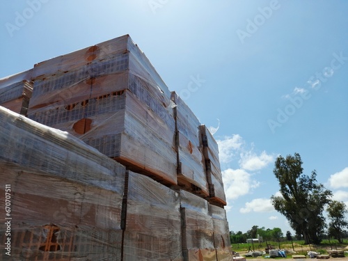 Pila de ladrillos huecos paletizados para la construcciòn de casas y edificios, protegidos con papel plàstico, en el corralón,  forma un original diseño con fondo de los árboles y el cielo azul
