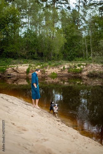 Man in Teal Poncho Standing by the River, Dog Playing by the Riverside