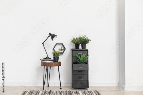 Desk lamp on small table and file cabinet with houseplants near white wall