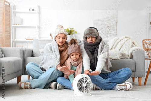 Frozen family in winter clothes warming near electric fan heater at home photo