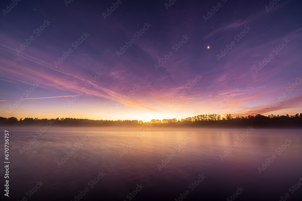 Obraz premium Sunrise at the quarry pond in Illingen. The sun's rays make the clouds glow while the stars still twinkle and fog drifts over the water.