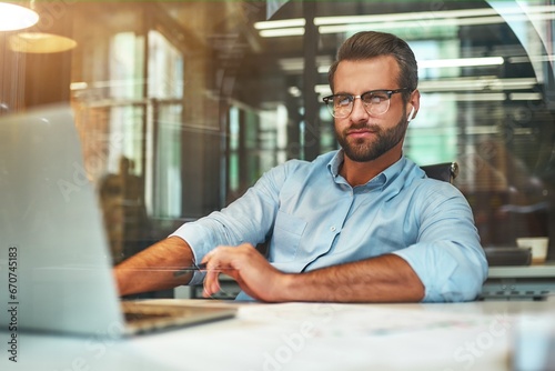 Successful businessman. Young bearded man in eyeglasses and formal wear working with laptop while sitting in the modern office
