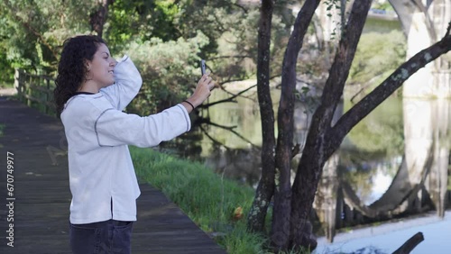 Young girl taking a selfie while grooming herself in a natural environment with water in the background. Brown curly hair with white sweatshirt. Concept of social networks and young people.